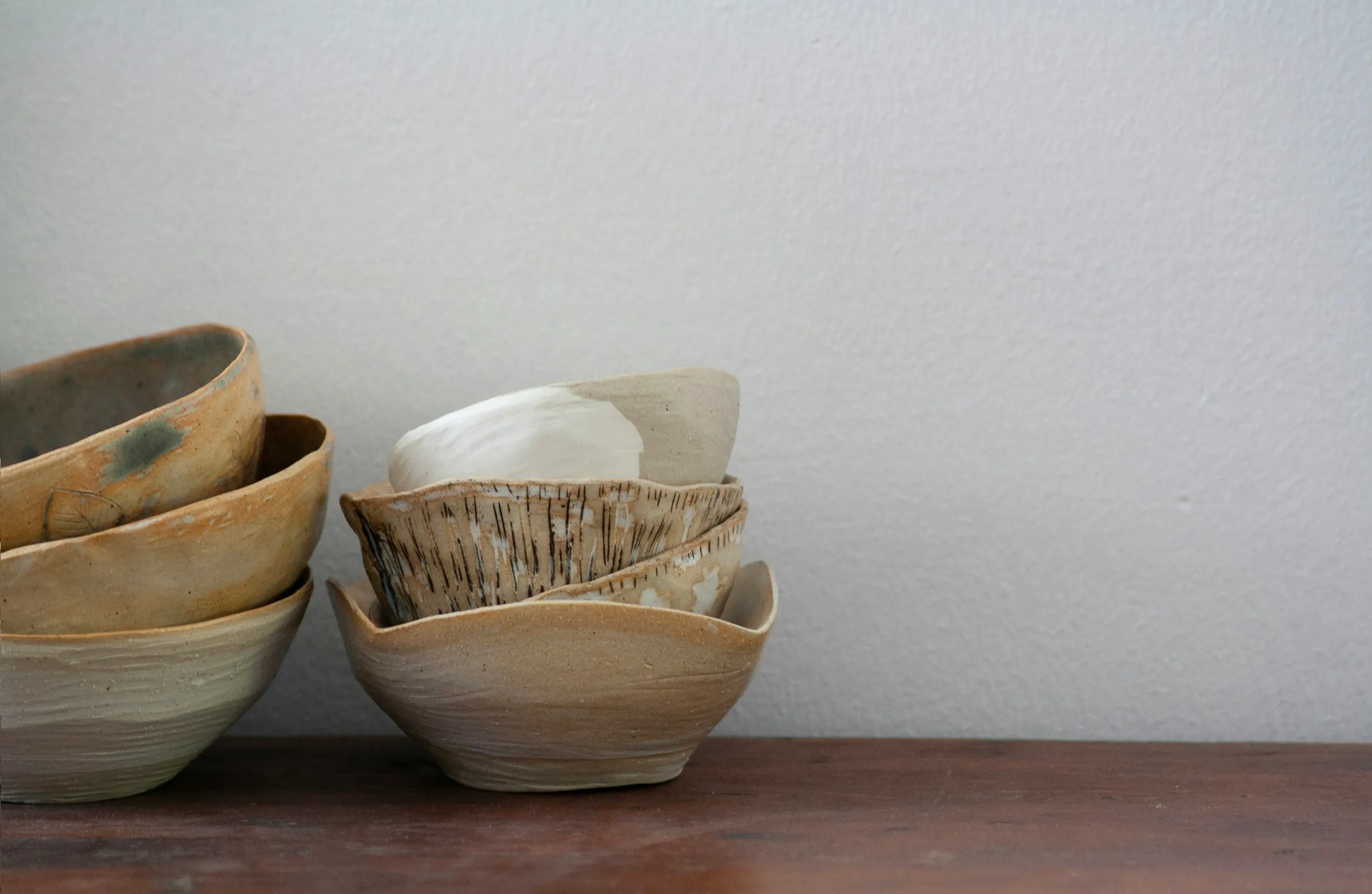 Stack of handmade ceramic bowls in sand and white tones on a wooden shelf against a white wall