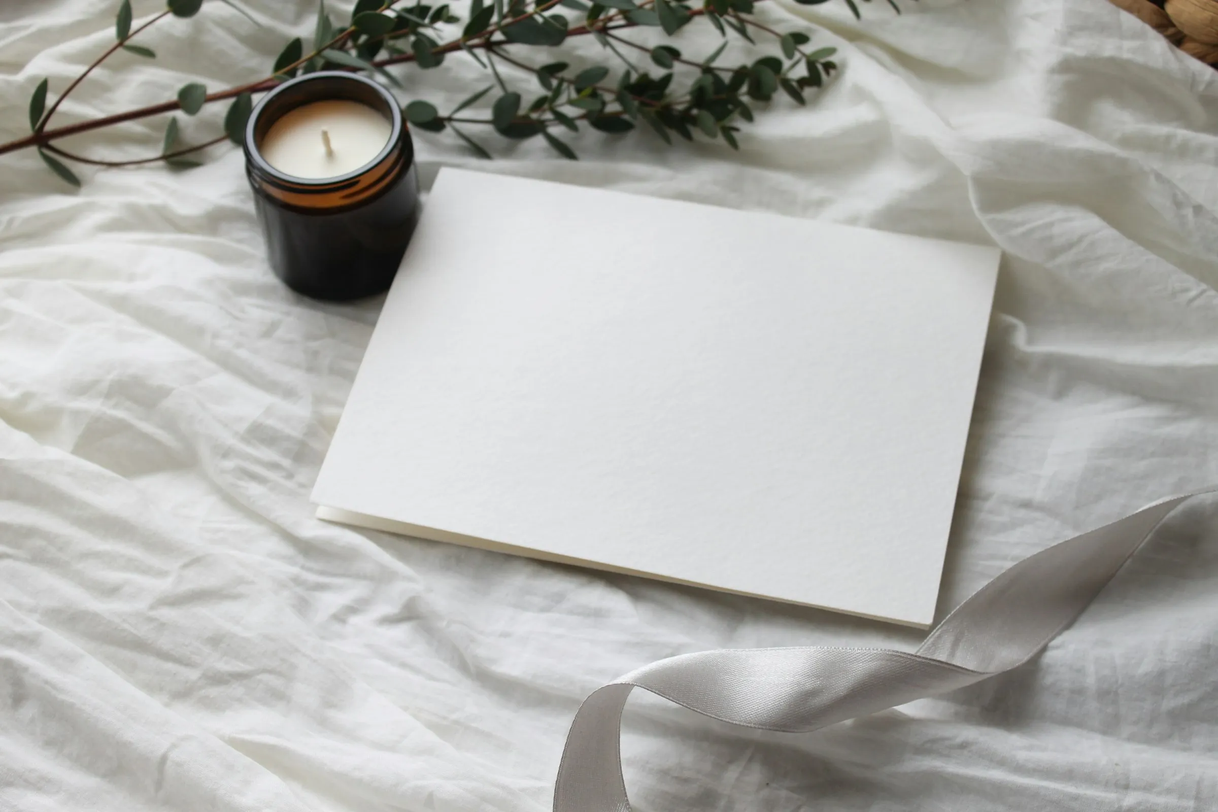 Small candle in a dark jar and a eucalyptus branch beside a blank card and white ribbon on crumpled white linen