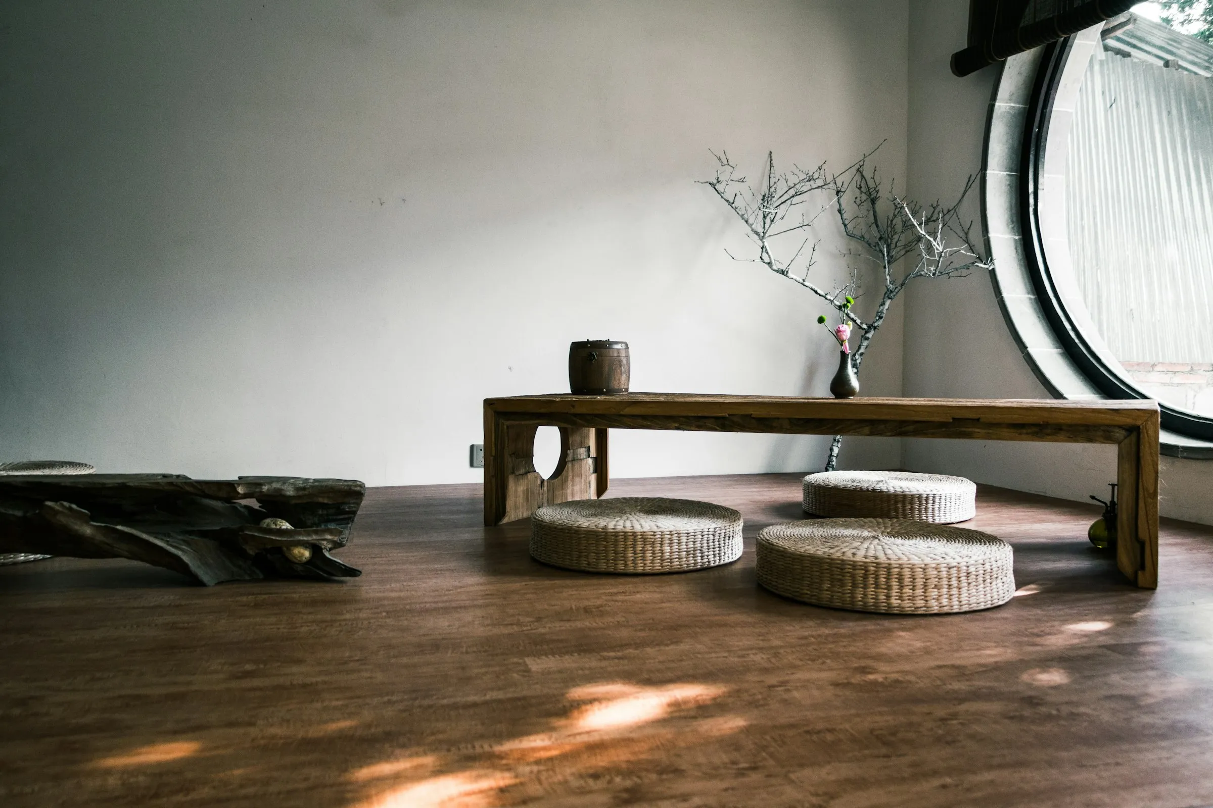 Low wooden table with woven straw floor cushions and a bare branch in a small vase, in a white room with a round window