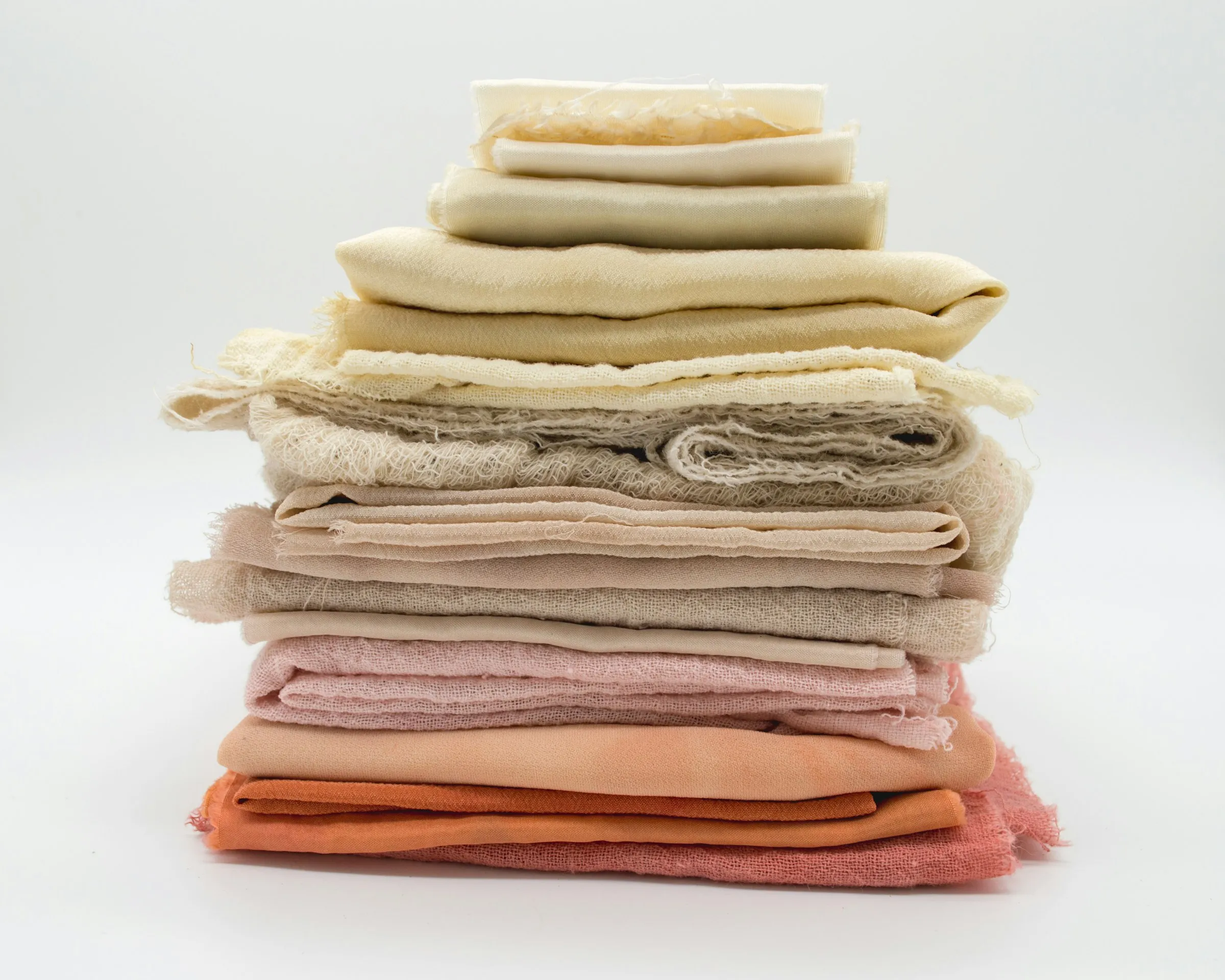 Stack of folded soft fabrics in cream, sand and peach tones on a white background